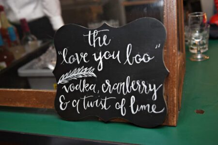Chalkboard sign reads: The love you loo vodka, cranberry & lime; on a bar counter with glasses and bottles in the background.