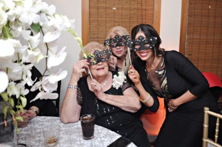 Three women in black with decorative masks smile at a festive indoor event; white flowers appear in the foreground.
