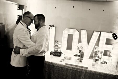 Two men in suits embrace by a glowing LOVE sign, sharing a romantic moment at a New Orleans wedding.