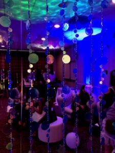 Blue and green beaded strings hang before a lounge with people on couches, lit by vibrant blue and purple lights.