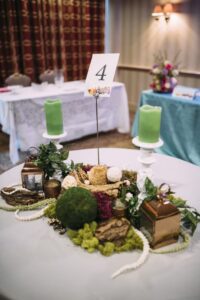 Round table with green candles, moss, bark, flowers, lanterns, and table number 4; other decorated tables in background.