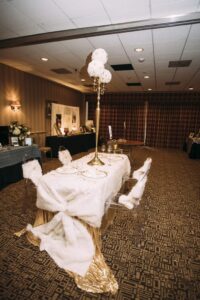 Elegant banquet table with gold sequins, white fur decor, gold plates, tall floral centerpiece, and clear chairs.