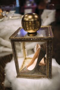 Sparkling high-heeled shoe in a beaded glass display box with gold sphere, set on a plush white surface.