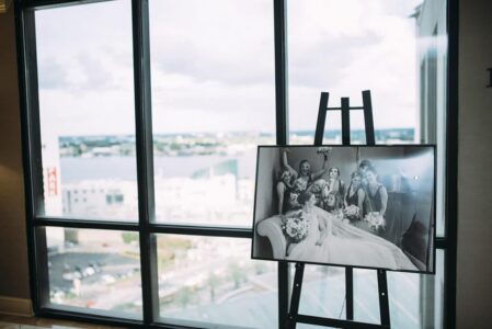 Black-and-white photo of women in dresses with bouquets on an easel before large windows showing a cityscape.