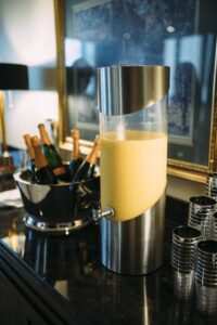 Orange juice dispenser beside champagne bottles and clear plastic cups on a dark countertop.