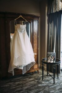 White strapless wedding dress on wooden wardrobe, with silver heels and New Orleans magazine on a table by gray-curtained window.