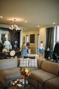 Warm modern living room with beige sectional, pillows, flower vase, and people chatting around tables.