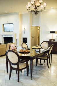 Modern dining area with four chairs, dark table set with food, chandelier, marble floor, beige sofa, TV, and fireplace.