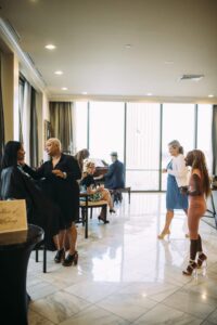 Makeup artist working on a woman as people chat and someone plays piano in a bright room with large windows and marble floors.