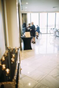 Guests in formal wear chat by a tall window in an elegant, bright room with marble floors; table with sign and candles in front.