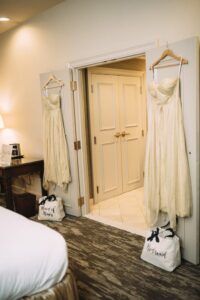 Cream dresses for Maid of Honor and Bridesmaid hang on doors in a neutral, carpeted bedroom with labeled bags below.