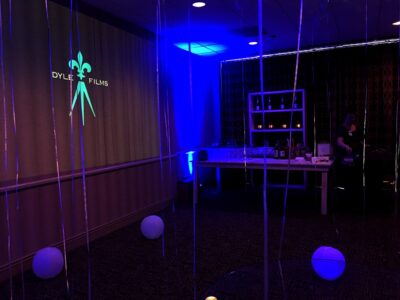 Blue-lit room with white balloons, streamers, DYLET FILMS projected on the wall, and a person near a snack table.