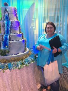 Smiling woman in blue top stands by large blue, white, and green cake, holding a folder and tote bag.