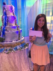 Smiling woman with envelope stands by tall decorated cake; colorful drapes and “Cake Pull for Prize” sign in background.