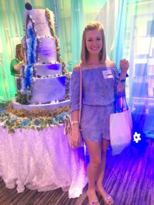 Smiling woman in a blue romper stands with a tote bag beside a decorated cake under blue and purple event lighting.