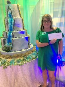 Woman in green dress smiles, holding envelope beside tall blue and white cake against colorful draped background.