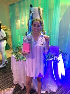 Woman in striped dress smiles, holding pink gift bag by tall decorated cake at party with green and blue decorations.