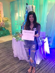 Woman with long dark hair in a gray tank top and ripped jeans holds a gift certificate indoors near a decorated cake and teal curtains.