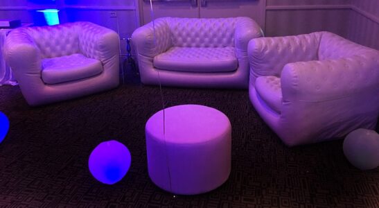 White tufted leather sofas and a round ottoman form a semi-circle on patterned carpet with balloons and soft purple lighting.