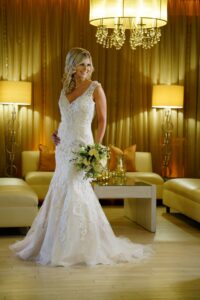 Bride in white lace gown smiling with bouquet in an elegant, gold-lit room featuring chandeliers and cream sofas.