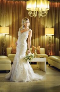 Bride in white lace gown with bouquet poses in elegant room featuring gold curtains, beige furniture, and warm chandelier lighting.
