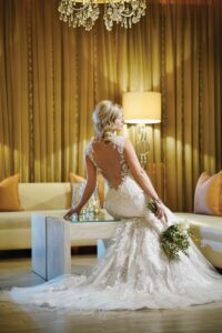 Bride in a lace wedding dress sits on a table, holding a bouquet, low back visible. Room has golden curtains and cream sofas.