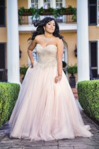 Woman in a strapless beaded ball gown poses outside a grand building with columns, greenery, and balconies.