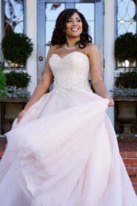 Woman in white strapless gown smiles outside, holding her dress, with a jeweled necklace, plants, and doorway behind her.