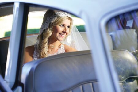 Smiling blonde bride in a lace veil and wedding dress sits in a classic car, gazing out the window.