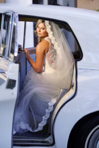 Bride in lace wedding dress and veil sits in classic white car, looking back with the door open.