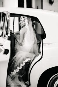Bride in lace veil sits in classic car’s backseat, looking over shoulder at camera. Black and white photo.