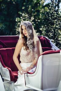 Bride in lace veil and white dress on red velvet seat in open white carriage, surrounded by greenery and sunlight.