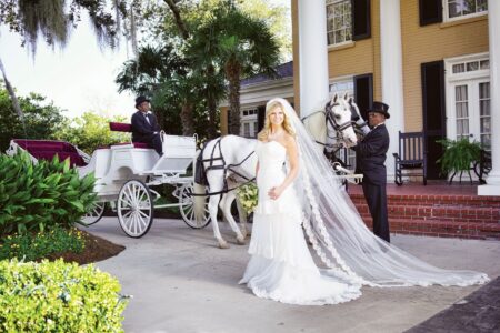 Bride in white dress and veil stands by white horse-drawn carriage, outside grand yellow house with columns and greenery.