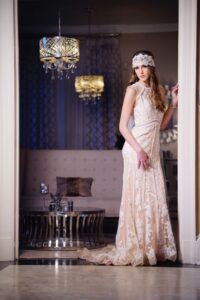 Woman in a lace dress and headpiece stands in a doorway, with a luxury living room and chandeliers behind her.