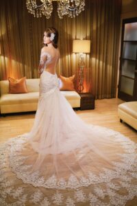 Bride in a long, lacy white wedding dress with dramatic train stands in elegant room with chandeliers and beige sofas.