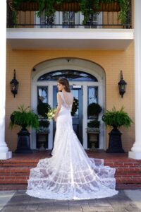 Bride in lace gown on brick steps, back to camera, by double doors with potted plants and greenery overhead.