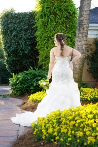 Bride in white gown on garden path with yellow flowers, holding bouquet and looking over shoulder amid lush green plants.