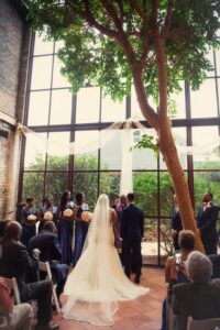 Bride and groom stand before an officiant under an indoor tree, surrounded by their wedding party and guests in a sunlit Louisiana venue.