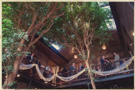 Formally dressed guests mingle on a light-filled, decorated balcony—an elegant New Orleans-inspired wedding scene.