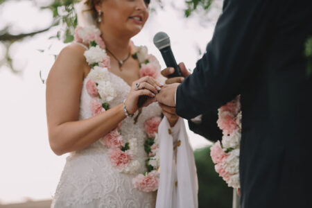 Bride in white dress puts ring on groom during outdoor wedding; both wear flower garlands, groom holds microphone.