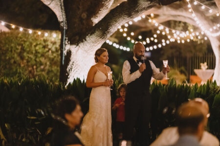Bride and groom toast with champagne under string lights at night, child between them, guests seated in front.