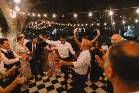 People in formal wear dance and celebrate under string lights on a checkered outdoor floor at night.