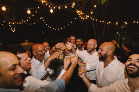 Men in shirts toast with drinks outdoors at night, smiling under string lights and trees.