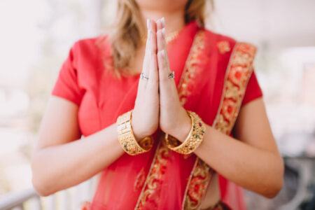 Woman in red and gold sari with gold bangles, hands pressed in prayer gesture; face not visible.
