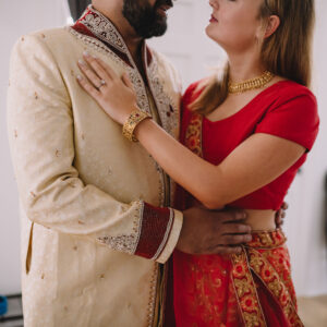 A couple in traditional Indian attire embraces closely; he wears a cream sherwani, she wears a red sari with gold jewelry.