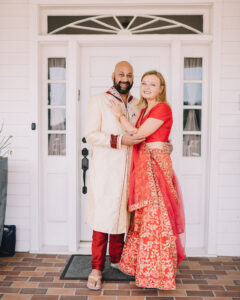 Smiling couple in traditional attire—man in cream and red sherwani, woman in red and gold saree—embracing by white door.