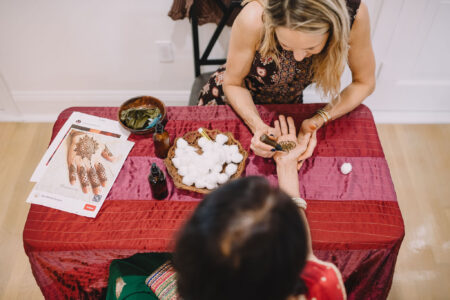 A woman applies henna to another’s hand at a red table with cotton balls, design sheets, and henna supplies visible.