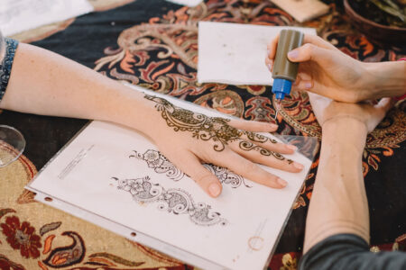 Someone applies henna designs to a hand with a reference sheet and patterned fabric in the background.