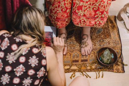 Applying intricate henna to feet in red embroidered clothing on an ornate rug; bowl of materials nearby.