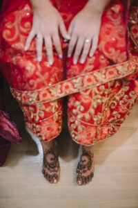 Person in red and gold attire sits with hands on lap, rings visible, white nails, and henna-decorated feet on wooden floor.